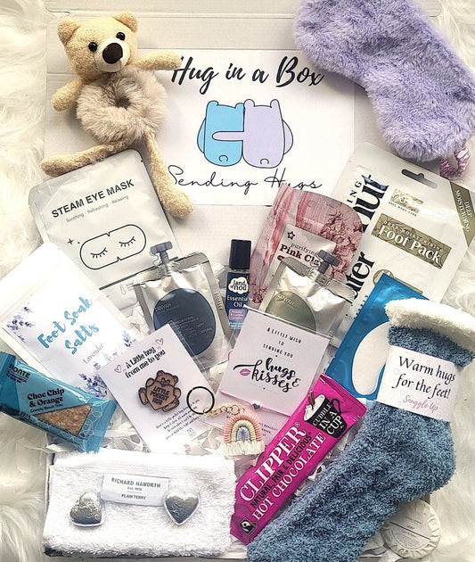 A variety of items spread out, including a teddy bear, socks, foot soak salts, a wish bracelet, a bear hug coin, hot chocolate, cookies, chocolates, a tea light candle, a rainbow keyring, and a personalized message card.